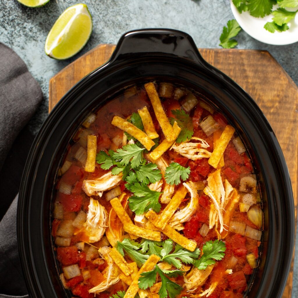 Chili in a slow cooker topped with shredded cheddar cheese, tortilla strips and chopped cilantro.