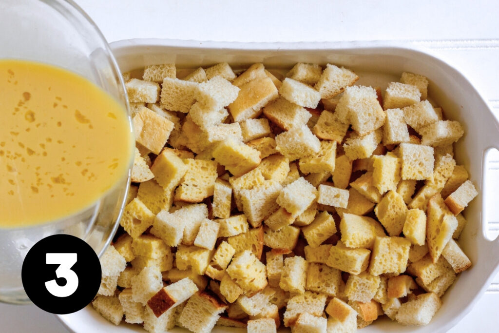 The egg mixture being poured over the cubed bread that is now in a casserole dish.