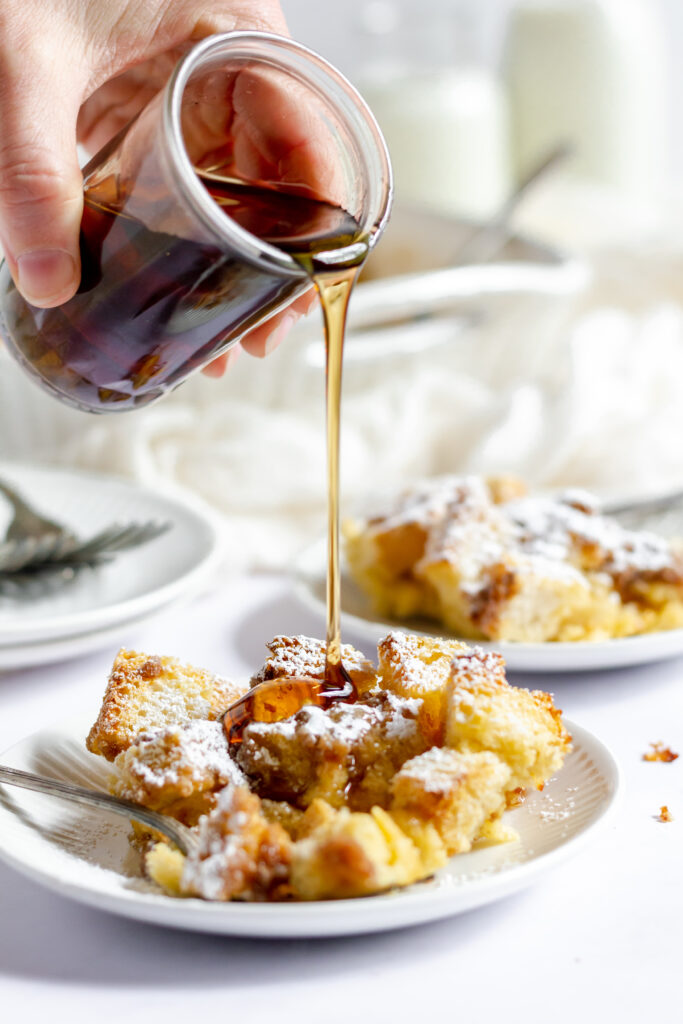 Someone pouring maple syrup over a plateful of French toast casserole that's been dusted with icing sugar.