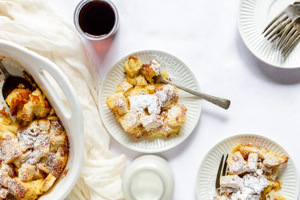 A casserole and 2 small plates of the baked French toast dusted with icing sugar. There is a cup of maple syrup beside.